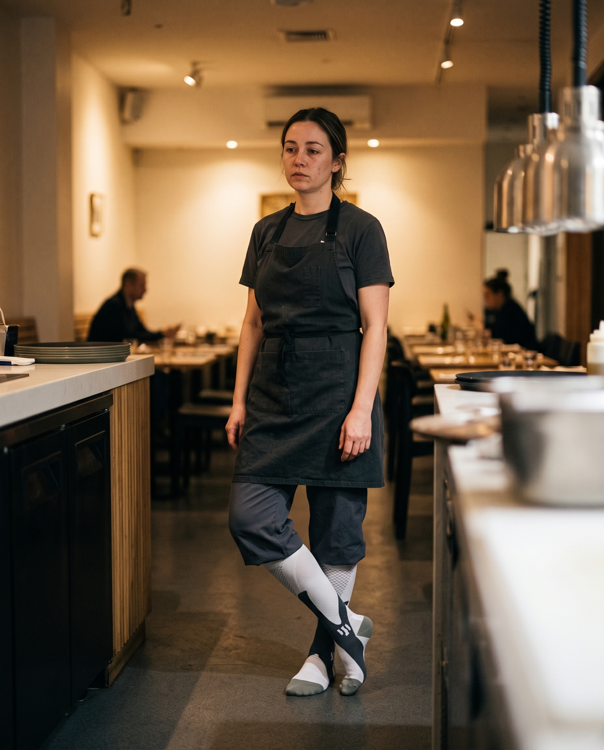 A hospitality worker standing behind the counter late in a long shift, wearing Montrell compression socks